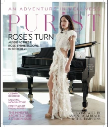 Rose Byrne wearing an ornate white ruffled dress standing beside a black grand piano in a sunlit interior space