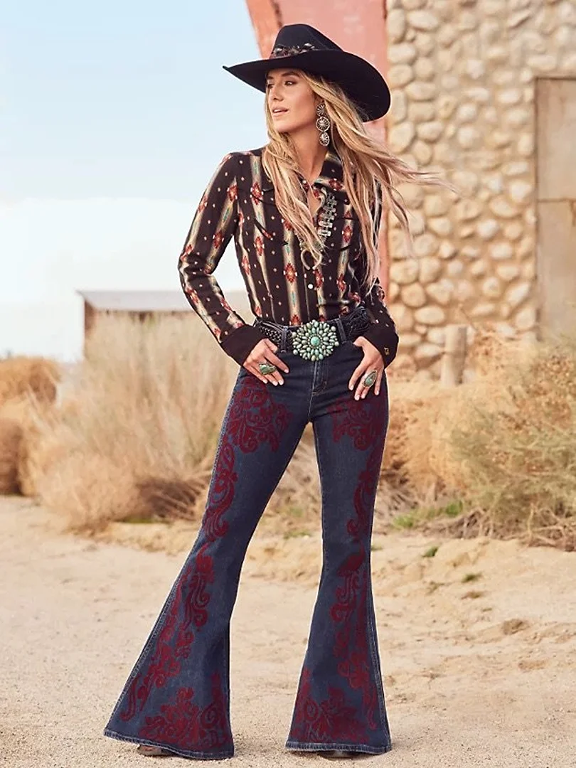 A woman in a cowboy hat and western shirt stands in a desert landscape, wearing denim bell bottoms with red embroidered details.