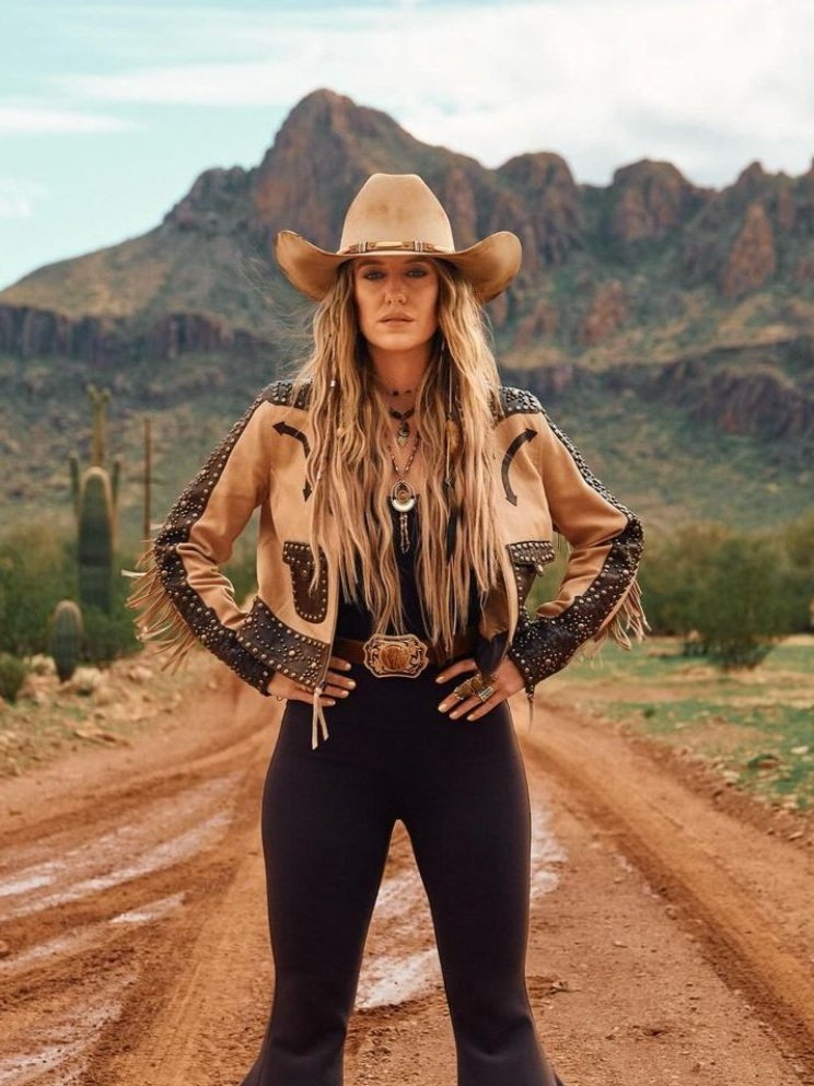 A woman wearing a tan cowboy hat and matching jacket poses confidently on a dirt road with a desert and mountain backdrop.