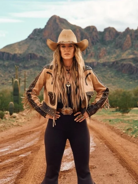 A woman wearing a tan cowboy hat and matching jacket poses confidently on a dirt road with a desert and mountain backdrop.