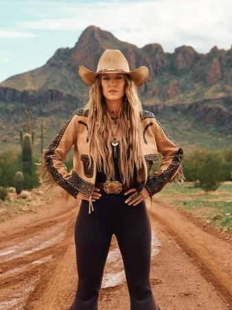 A woman wearing a tan cowboy hat and matching jacket poses confidently on a dirt road with a desert and mountain backdrop.