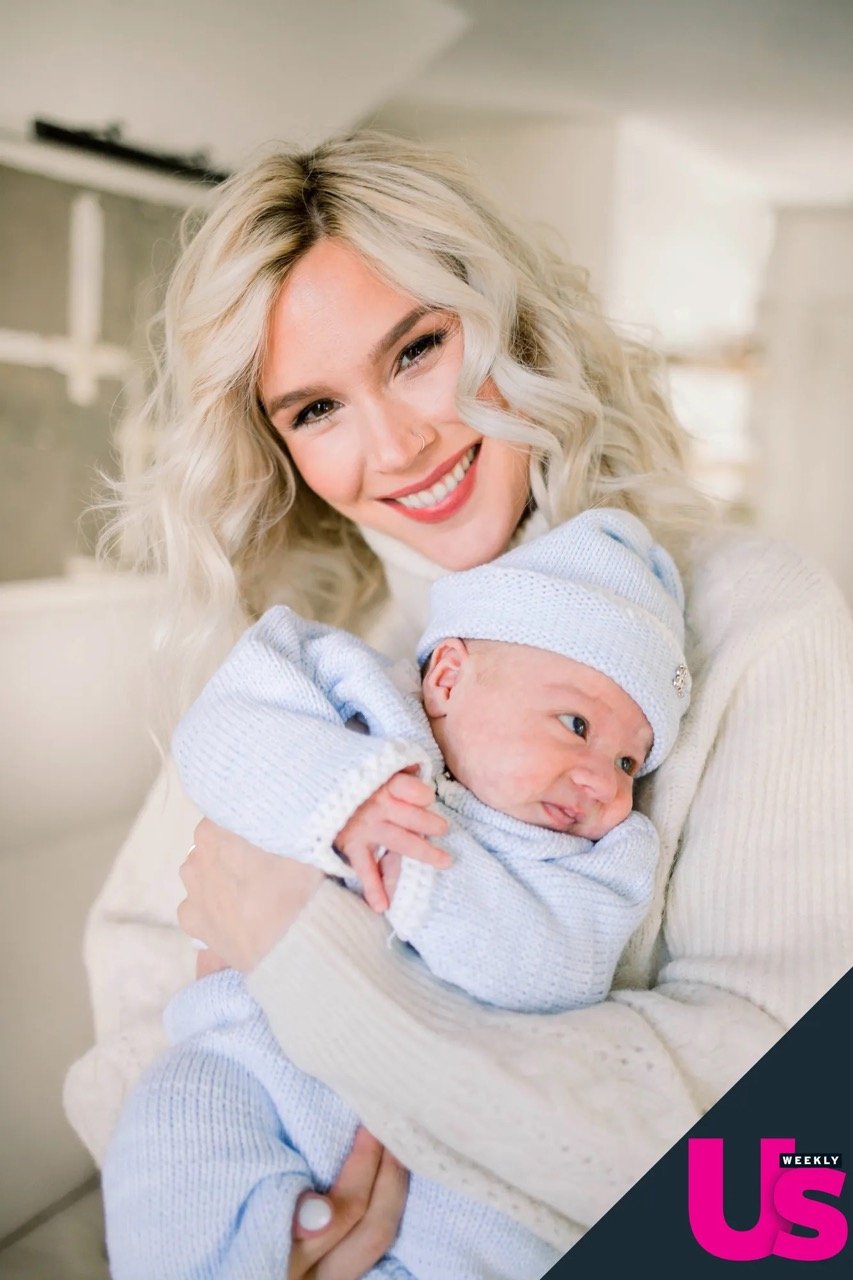 Smiling woman in a white turtleneck sweater holding a newborn baby in a blue knitted outfit in an indoor setting.
