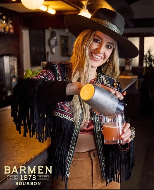 Blonde woman in a hat and fringed jacket pours a drink from a metal shaker into a glass in a dimly lit bar setting.