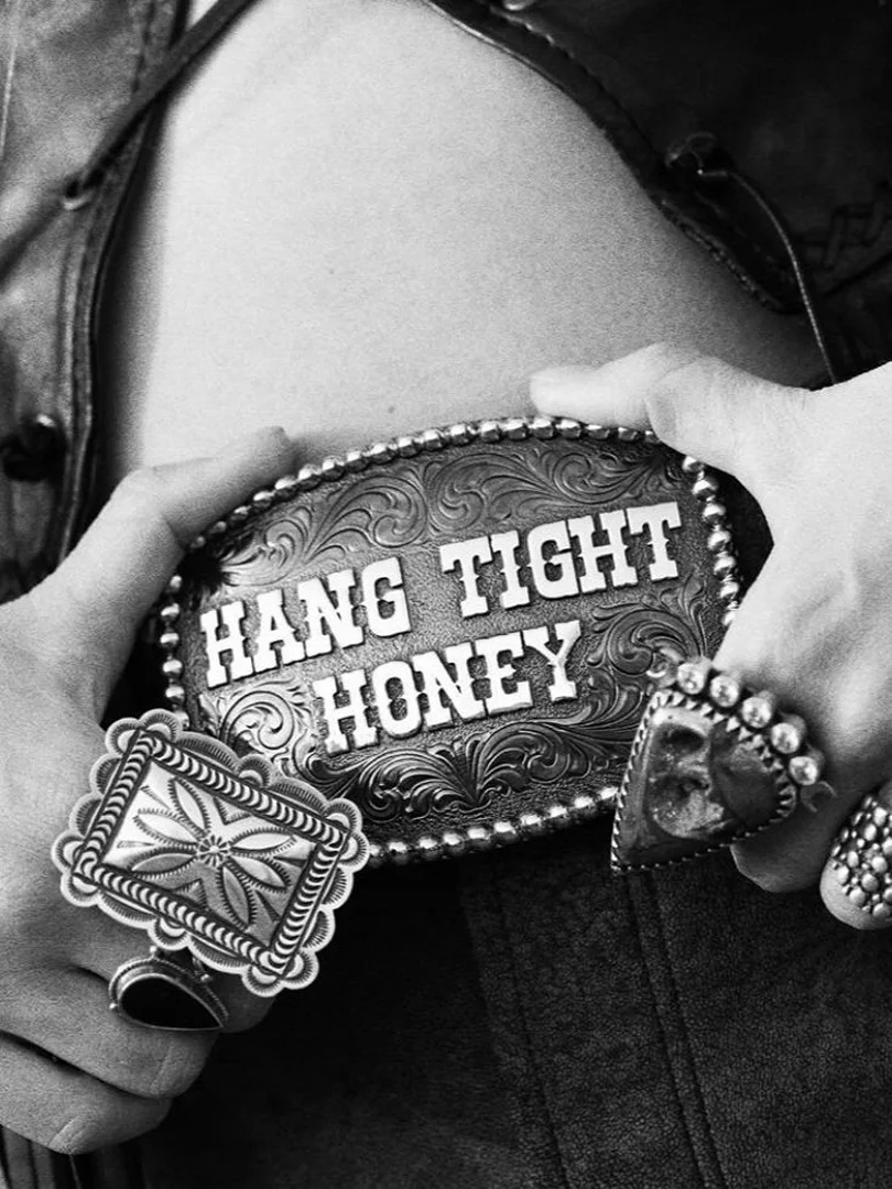 Close-up shot of a person's hands holding a decorative belt buckle that reads 'Hang Tight Honey,' accessorized with multiple statement rings.