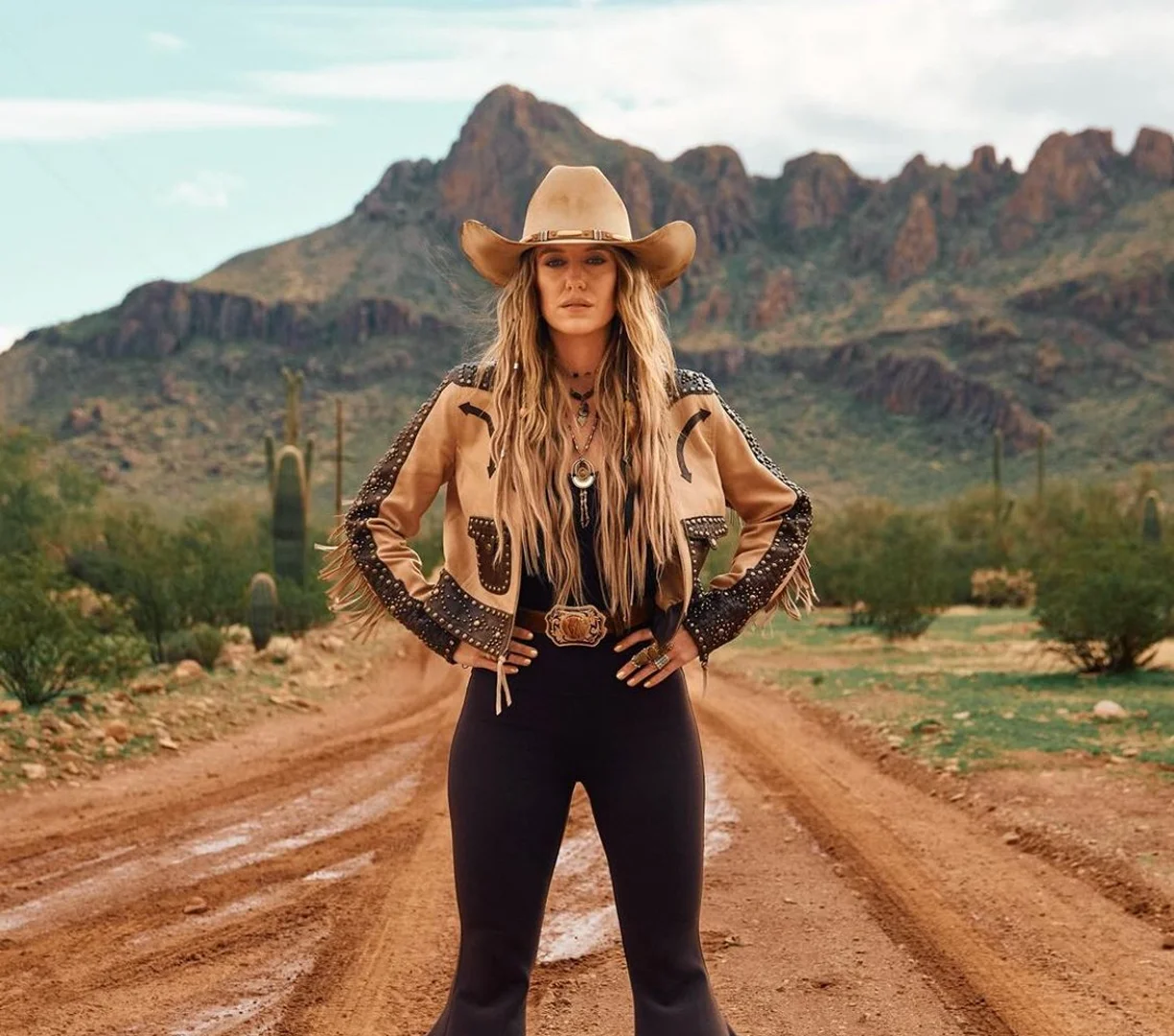 Woman in cowboy hat and jacket poses on dirt road with mountains in background, creating a Western-themed fashion shot.
