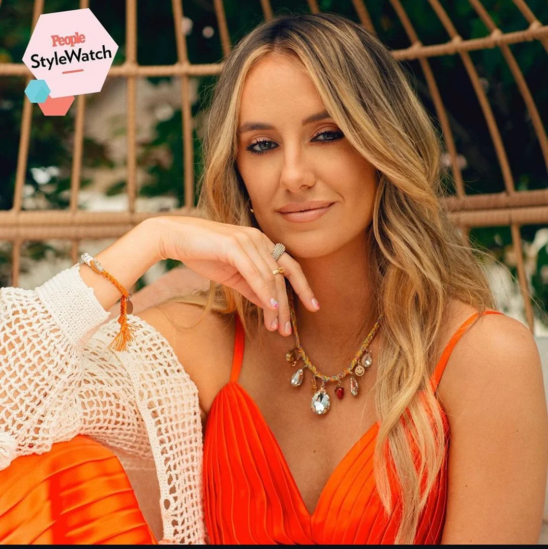 Portrait of a smiling woman with blond hair wearing an orange dress and jewelry posing in front of a leafy green background.