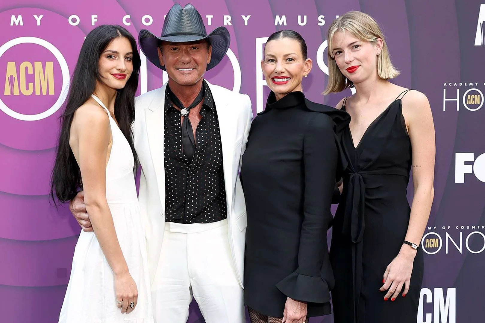 Group of four people on a red carpet. A man in a cowboy hat stands with three women in dark and light colored dresses.