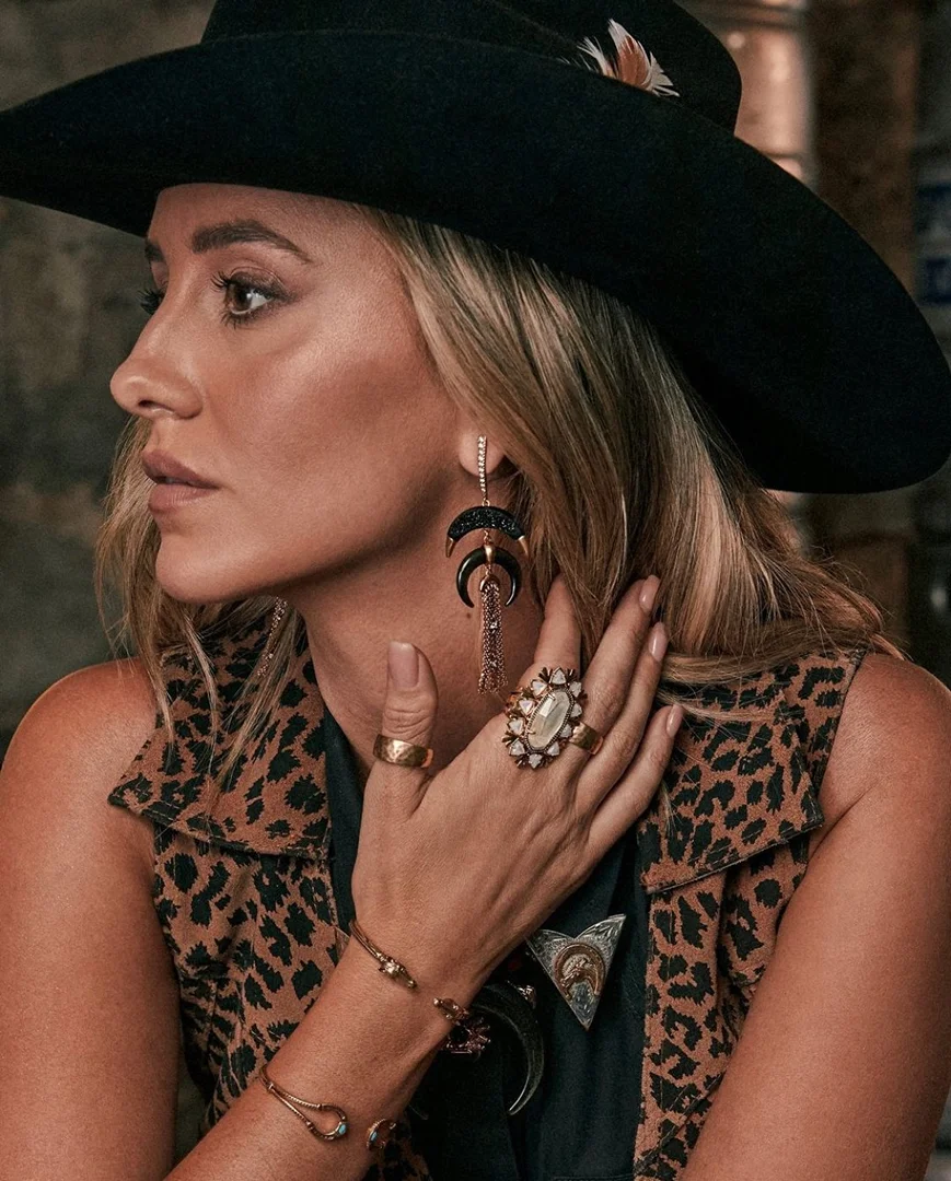 Portrait of a blonde woman in a black hat and leopard print vest, adorned with large earrings and rings, posing indoors.