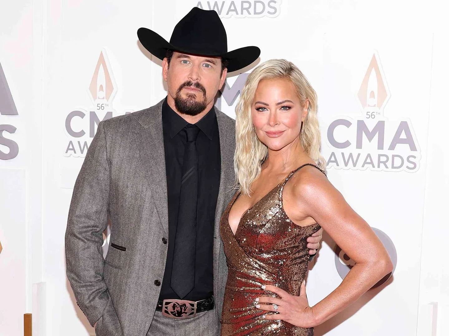Singer couple, Kellie Pickler and Kyle Jacobs, stand at the 56th CMA Awards wearing a black cowboy hat, grey suit, and gold dress.