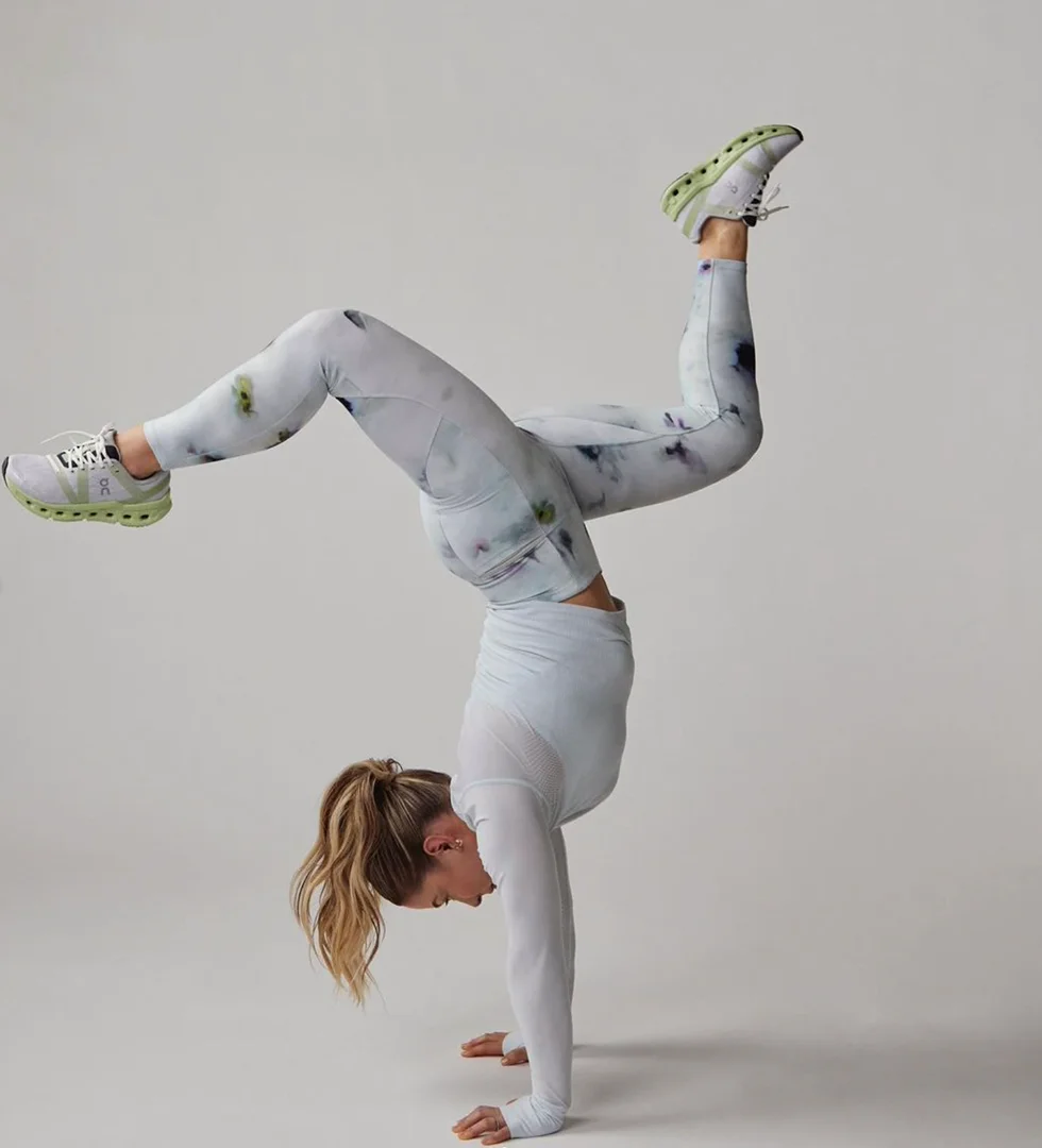 A woman in athletic wear performs a handstand with legs split in a bright studio setting, showcasing fitness and flexibility against a gray background.