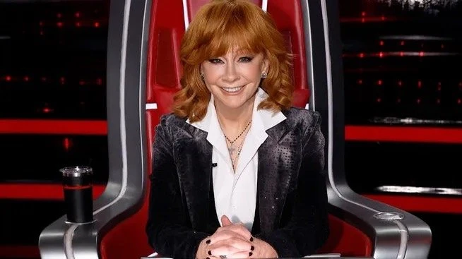 Reba McEntire sits in a red chair, smiling in a TV studio setting, wearing a black blazer and white top, hands clasped in front of her.