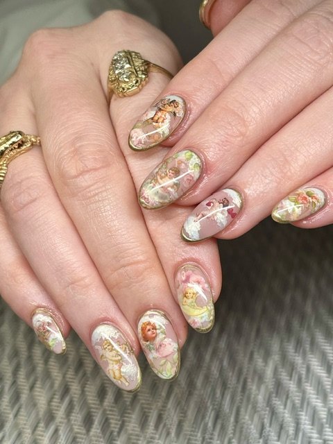 Close-up of a manicured hand featuring nail art with cherub and floral designs, adorned with gold rings, against a neutral backdrop.