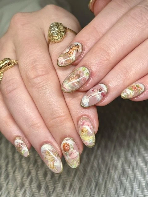 Close-up of a manicured hand featuring nail art with cherub and floral designs, adorned with gold rings, against a neutral backdrop.
