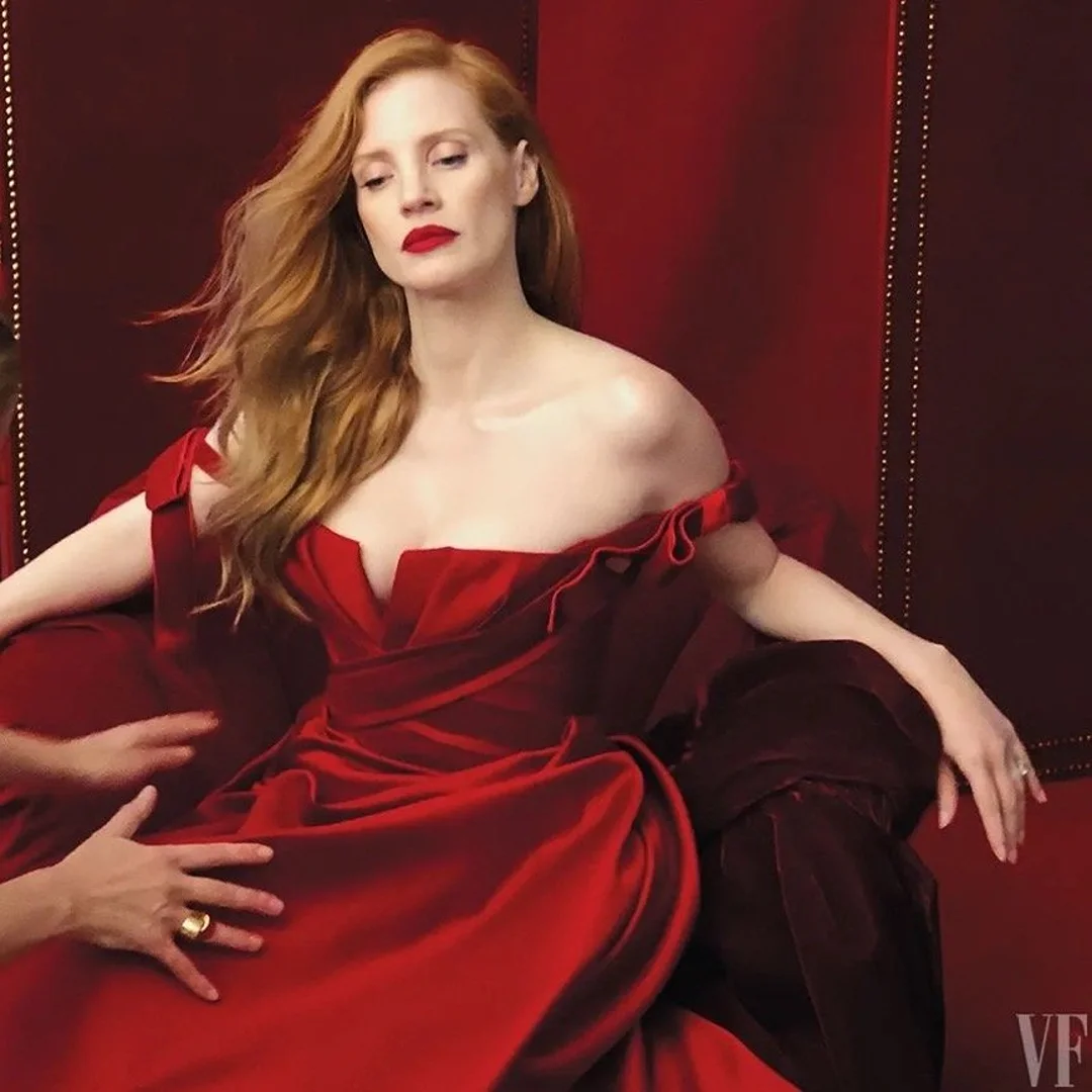 Elegant Jessica Chastain poses in a red dress on a matching red velvet chair, hands adjusting her gown in a studio setting.