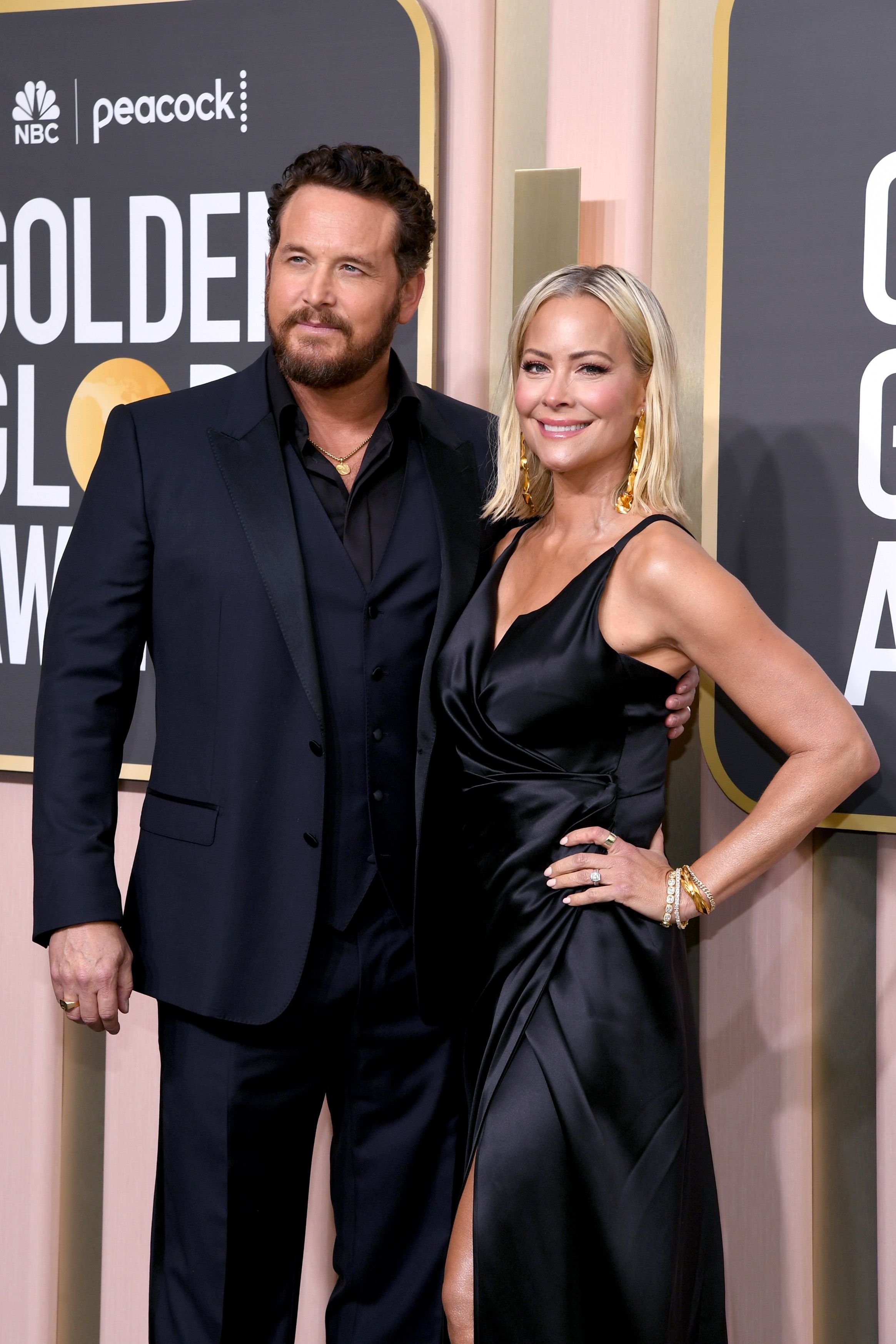 Man in suit and woman in black dress posing on red carpet at event. Golden Globes signage visible in the background. 