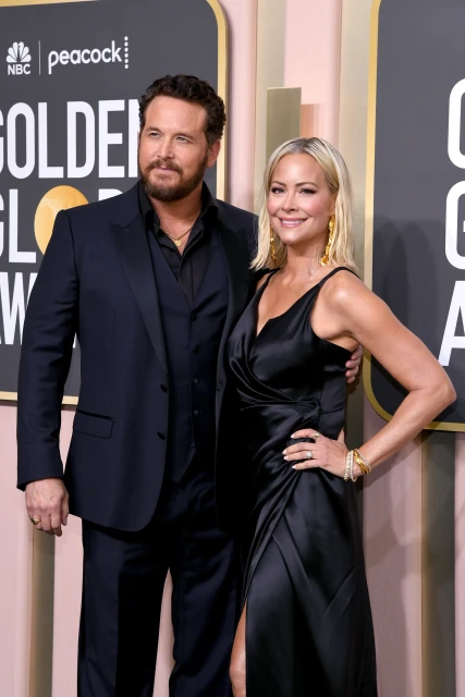 Man in suit and woman in black dress posing on red carpet at event. Golden Globes signage visible in the background. 