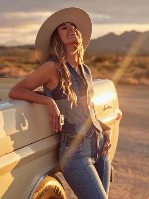 A smiling woman wearing a hat and denim leaning on a vintage truck in a desert landscape with mountains in the background at sunset.