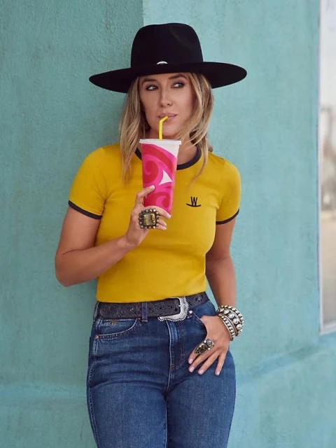 Blonde woman with a black hat drinking from a paper cup with a straw wearing a yellow and black shirt, jeans, and rings outside.