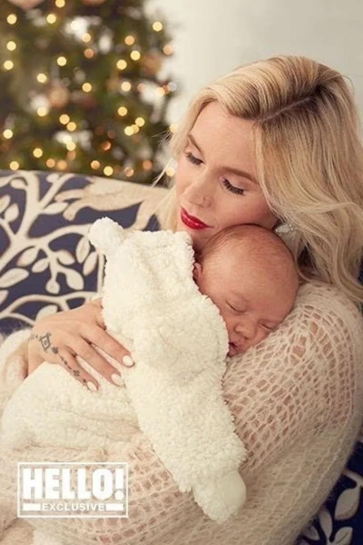 Woman with blonde hair in a beige sweater holds a sleeping baby in a white blanket with Christmas tree in the background.