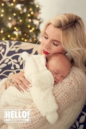 Woman with blonde hair in a beige sweater holds a sleeping baby in a white blanket with Christmas tree in the background.