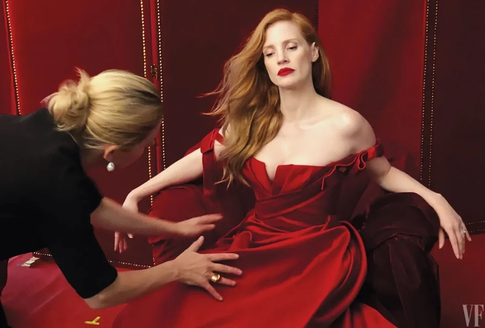 Jessica Chastain poses in a red off-shoulder dress while a stylist adjusts her gown in an indoor studio with a red backdrop.