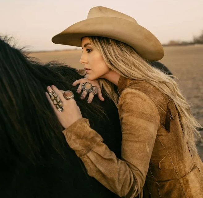 Woman in a tan cowboy hat and brown suede jacket leaning on a black horse in a tan-colored field at dusk, wearing rings on her fingers.
