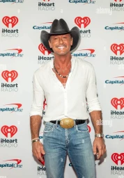 Man wearing a black cowboy hat, white buttoned shirt, and blue jeans standing in front of a branded step and repeat backdrop.