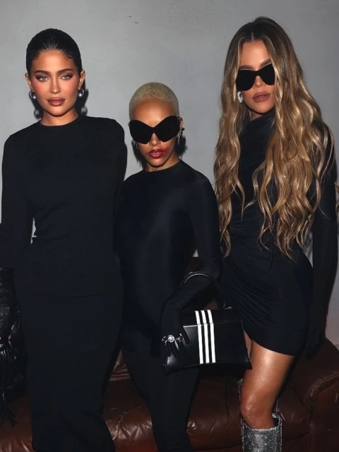 Three adult women pose indoors, wearing dark dresses and sunglasses; one carries a black-and-white bag with vertical stripes.
