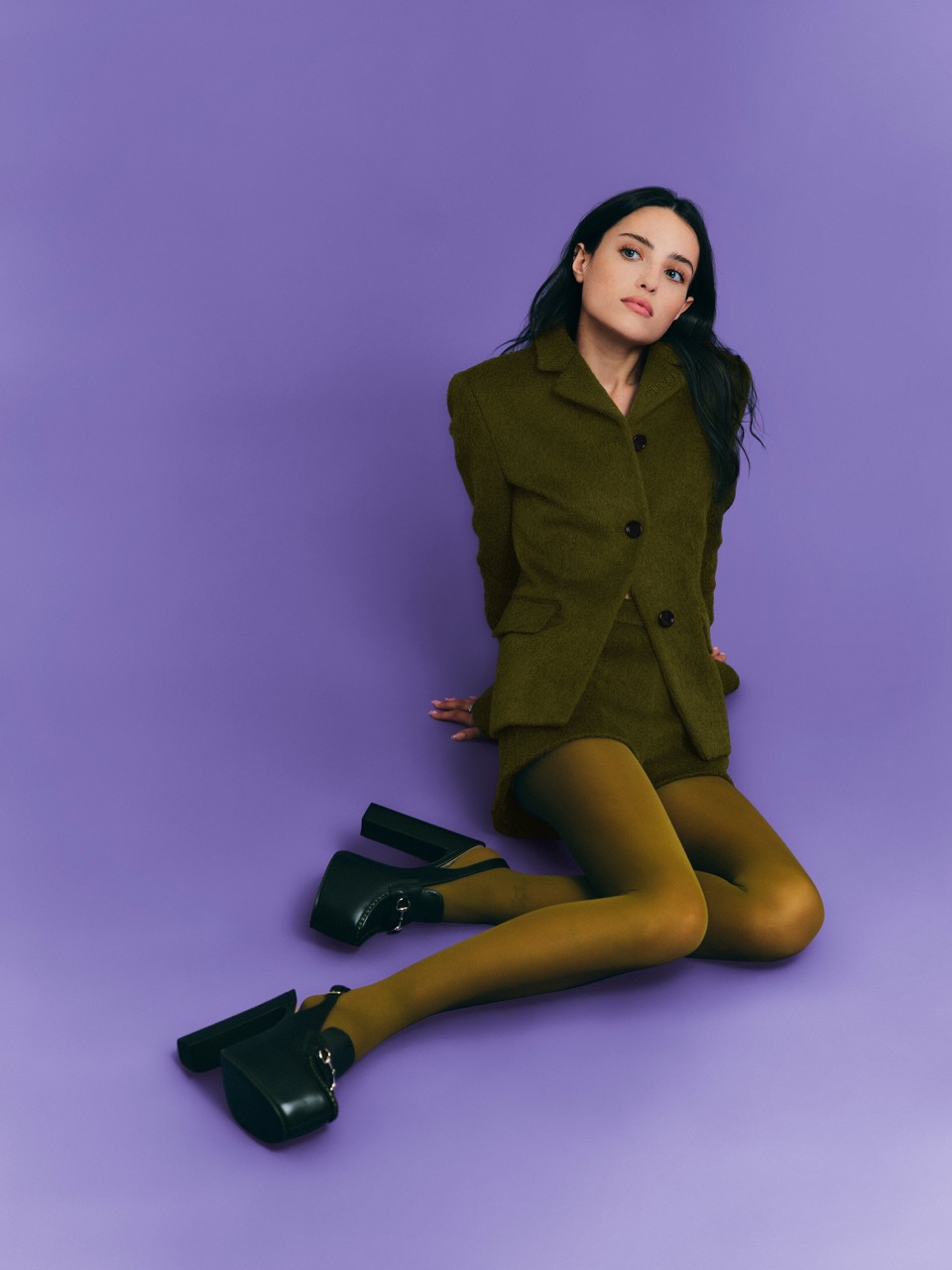 A young woman with long dark hair sits on a lilac-colored floor, wearing a green tailored suit with matching tights and chunky black heels.