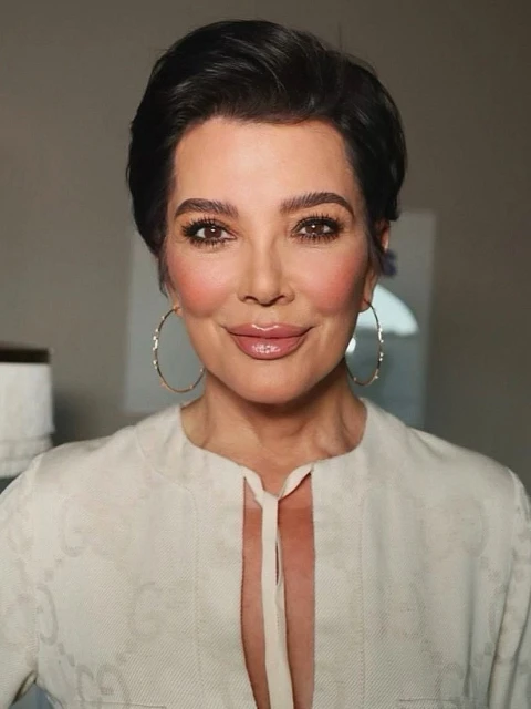 Portrait of Kris Jenner with short dark hair, gold hoop earrings, and a cream-colored top with a white background in a studio.