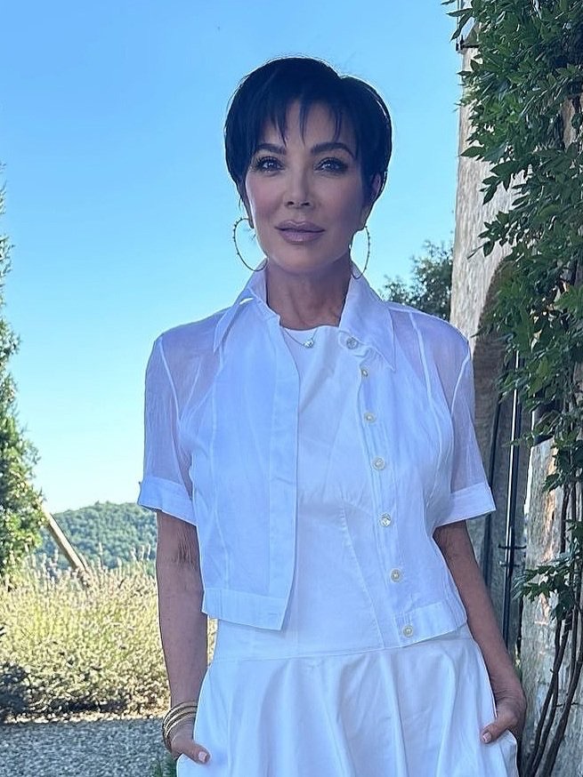 Close-up portrait of Kris Jenner wearing a white outfit, large hoop earrings, and standing outdoors with trees and a blue sky in the background.