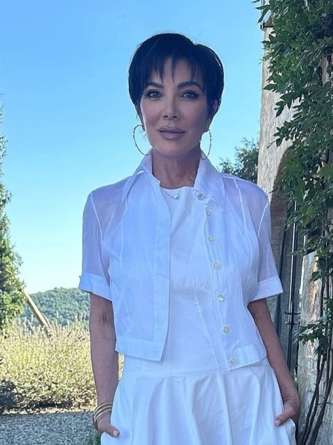 Close-up portrait of Kris Jenner wearing a white outfit, large hoop earrings, and standing outdoors with trees and a blue sky in the background.