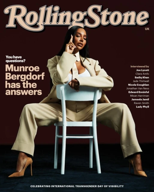 Munroe Bergdorf sits on a white chair for Rolling Stone UK cover. Wearing a tan suit, top, and heels. She has long dark hair.