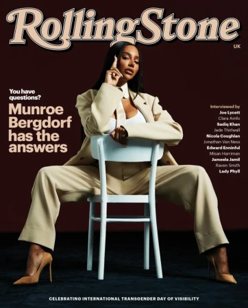 Munroe Bergdorf sits on a white chair for Rolling Stone UK cover. Wearing a tan suit, top, and heels. She has long dark hair.
