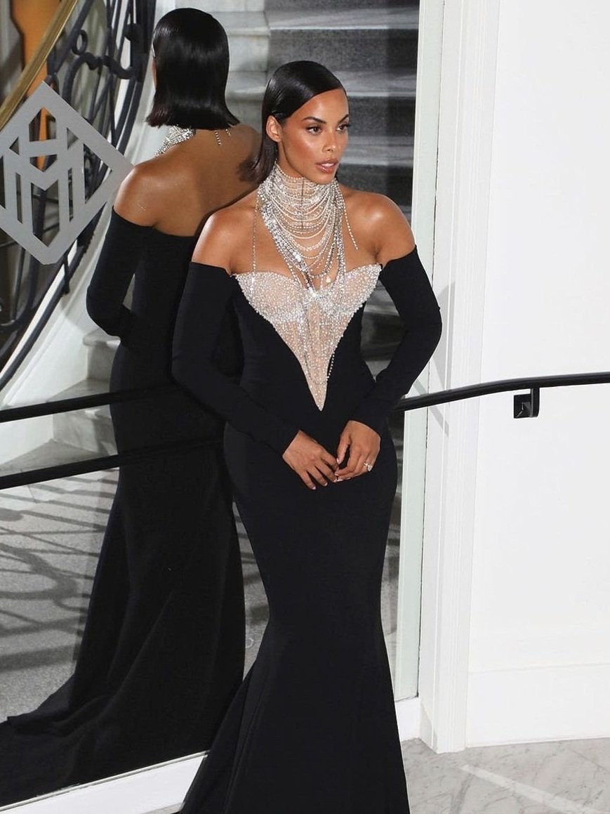 Elegant woman in a long black gown poses with jewelry, reflecting in a mirror, showcasing sophistication and style.