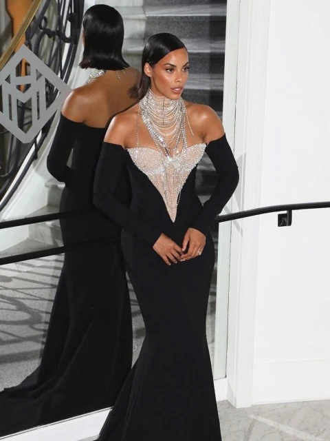 Elegant woman in a long black gown poses with jewelry, reflecting in a mirror, showcasing sophistication and style.