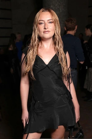 Woman with long wet-look blonde hair wearing a black sleeveless minidress standing indoors in London