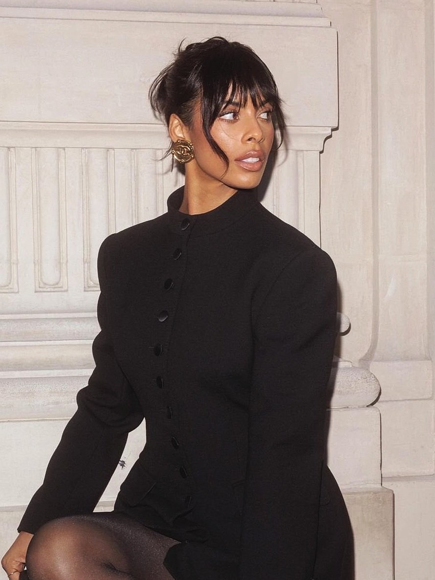 Elegant portrait of a woman in a black jacket and dark tights, sitting against a textured off-white wall with classic architectural details.
