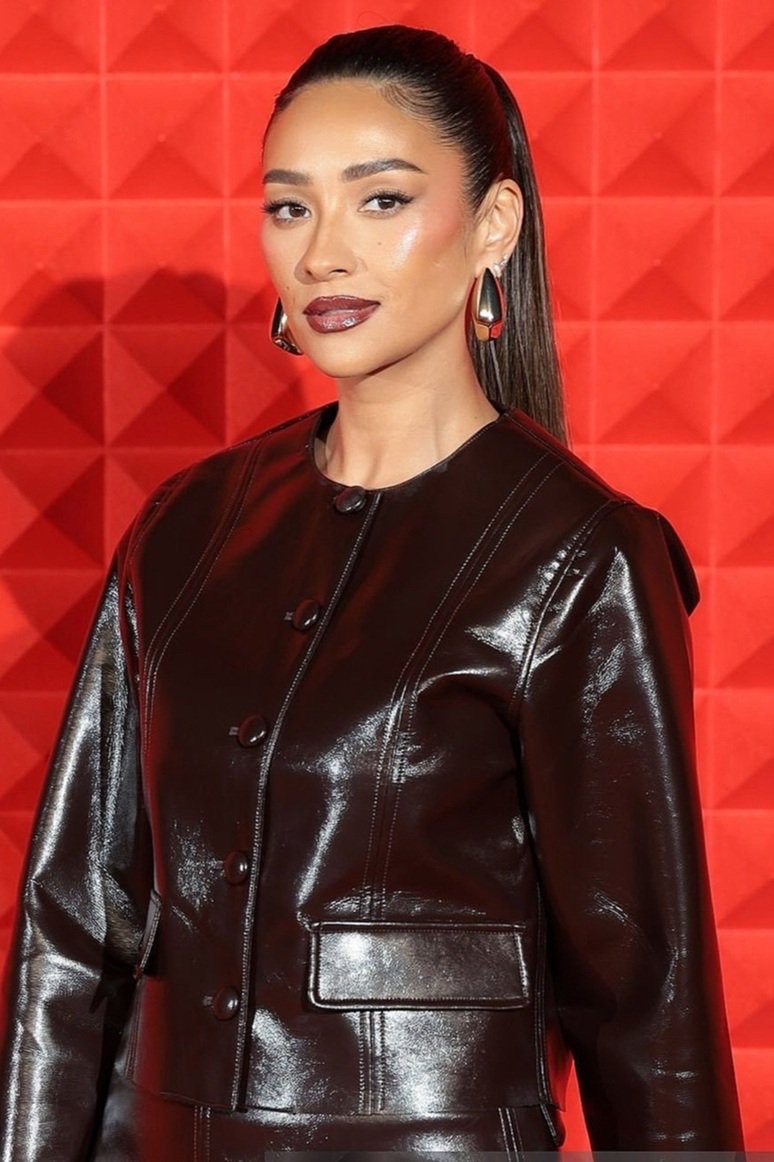 Portrait of a woman with brown hair in a high ponytail, wearing a shiny brown jacket and statement earrings, set against a red geometric background.
