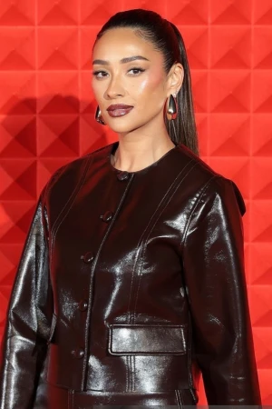 Portrait of a woman with brown hair in a high ponytail, wearing a shiny brown jacket and statement earrings, set against a red geometric background.