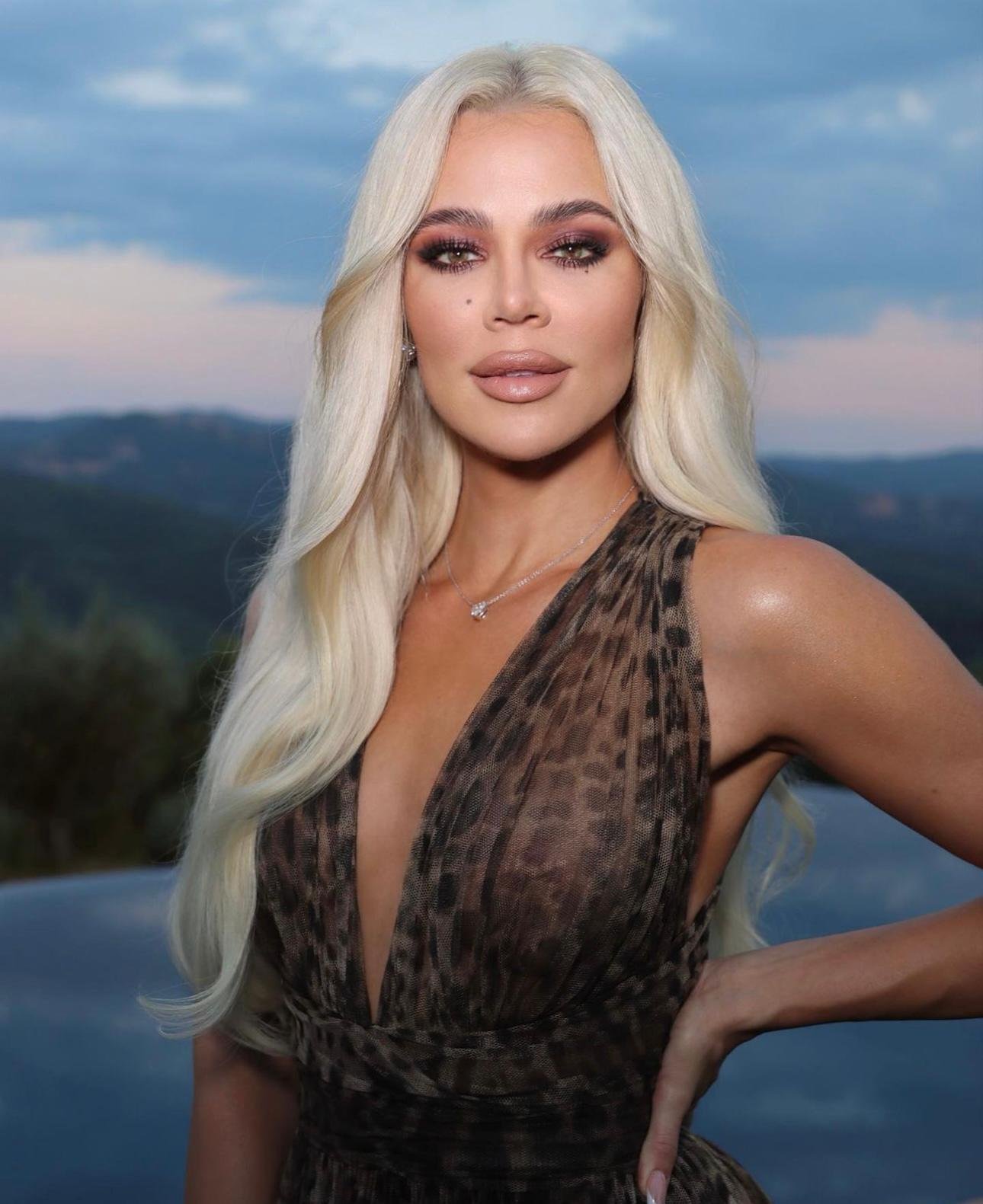 Close-up of Khloe Kardashian with long blonde hair wearing a leopard print dress, standing in front of a mountain landscape at dusk.