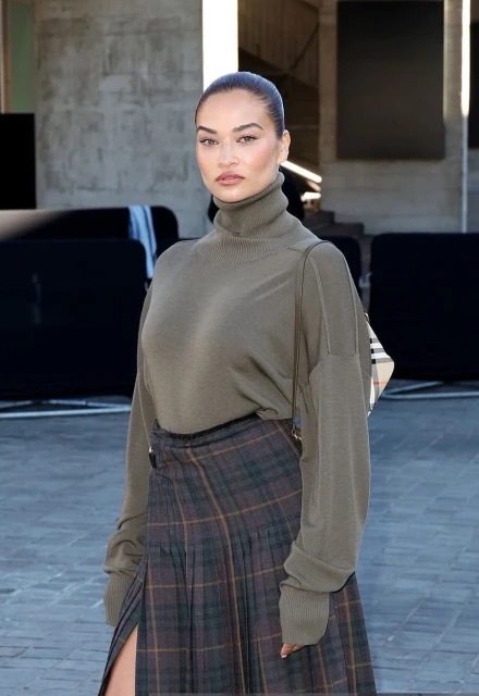 A woman poses in an olive green turtleneck and plaid skirt with a slit, accessorized with a handbag, in an urban setting.