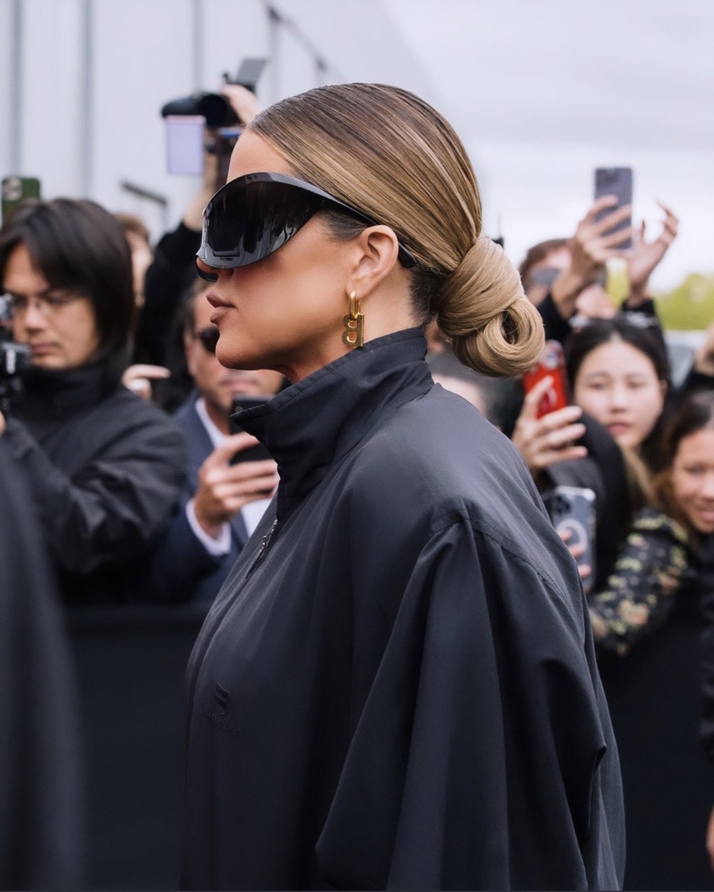 Woman with slicked-back hair and visor sunglasses in a crowd of people holding up phones to take photos in an outdoor setting.