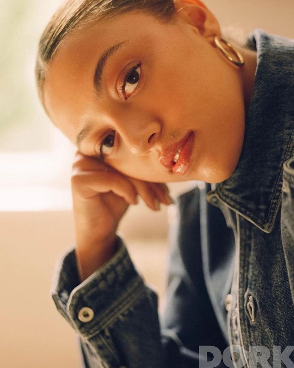 Close-up portrait of a young adult woman with gold hoop earrings wearing a denim jacket, resting her head on her hand.
