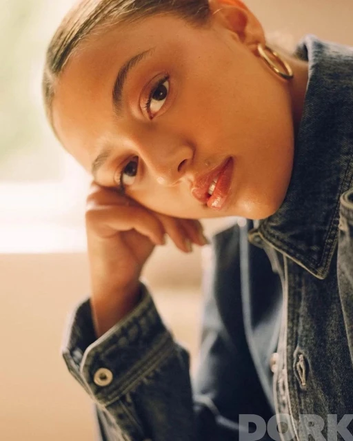 Close-up portrait of a young adult woman with gold hoop earrings wearing a denim jacket, resting her head on her hand.