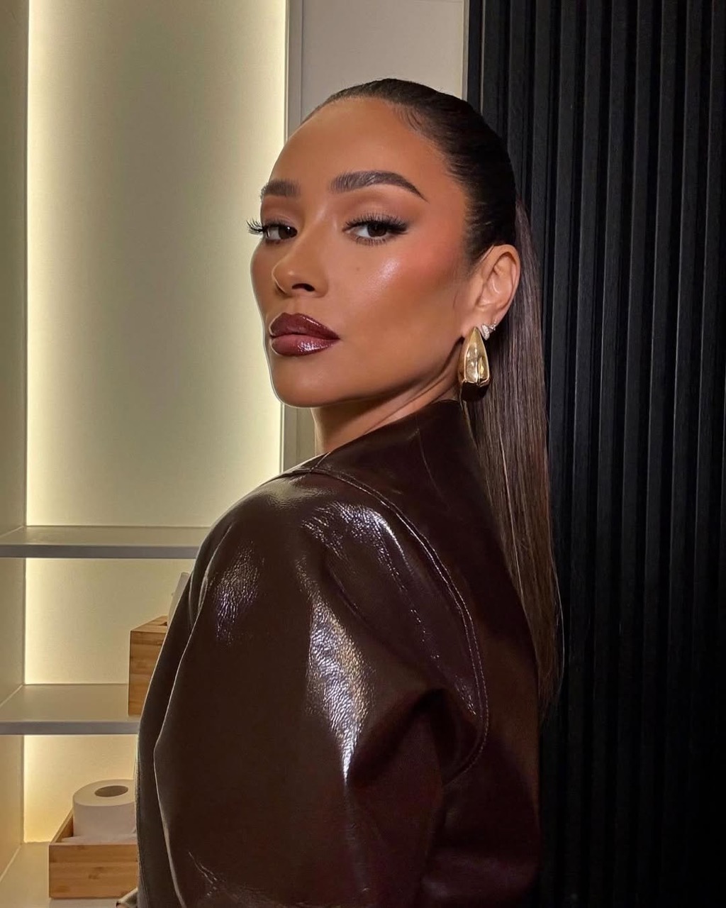 Portrait of a poised woman with sleek hair, glossy makeup, and a brown patent leather jacket, set against a modern interior backdrop.