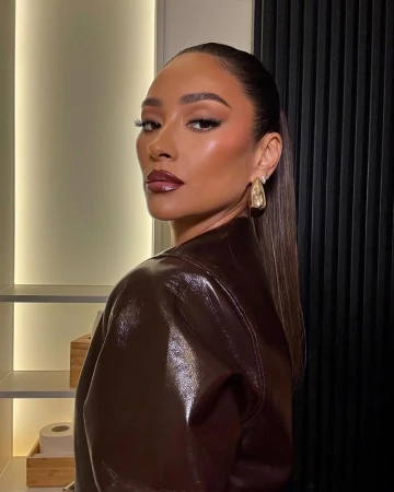 Portrait of a poised woman with sleek hair, glossy makeup, and a brown patent leather jacket, set against a modern interior backdrop.