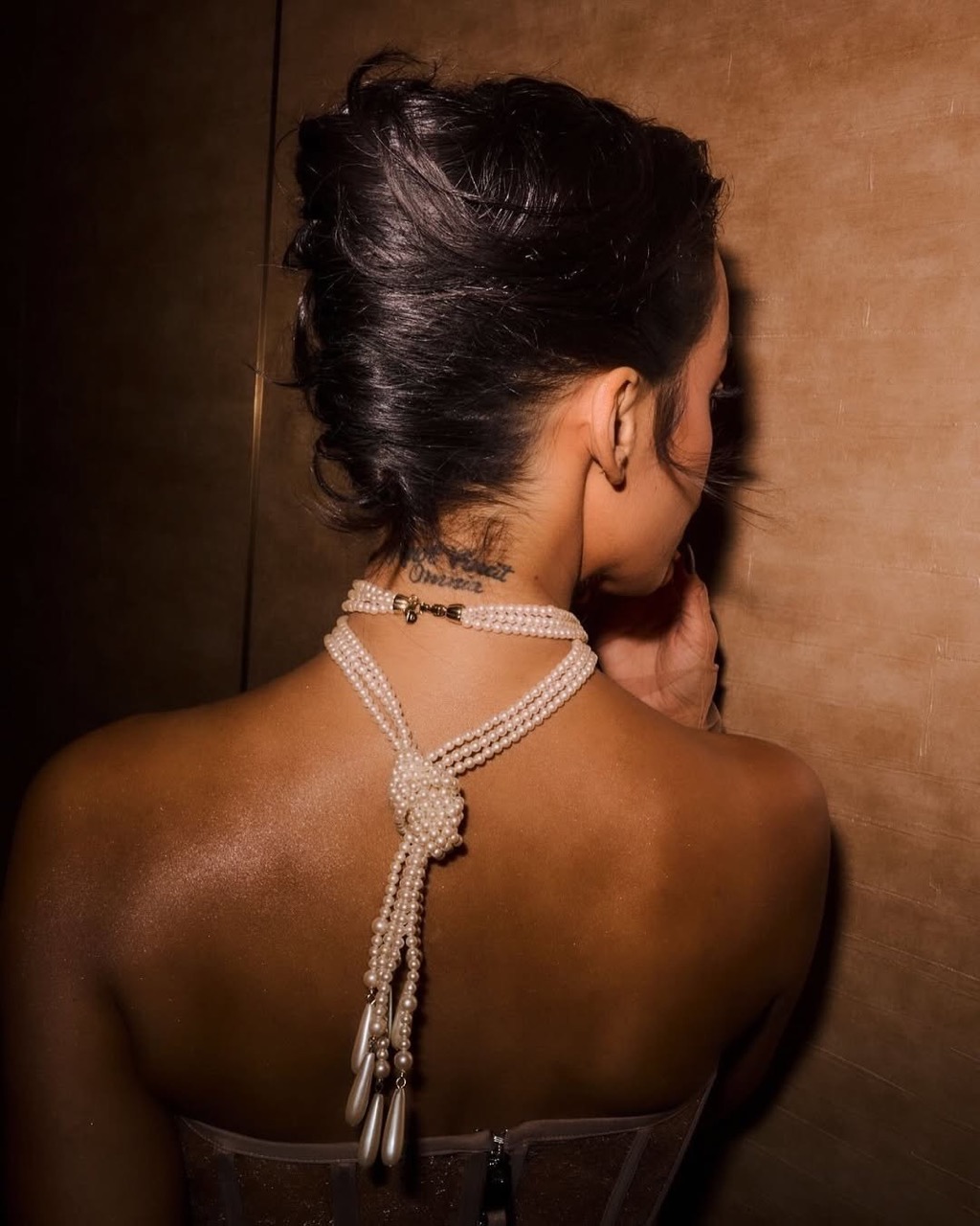 Elegant woman's back view showcases her updo, pearl necklace, and corset in a dimly lit setting, exuding classic glamour and sophistication.