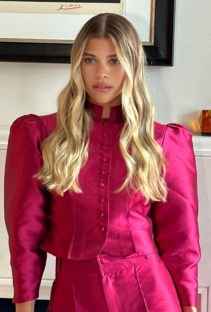 A blonde woman in a pink blazer and skirt is centered in a bright indoor shot, showcasing fashion and style with artwork in the background.