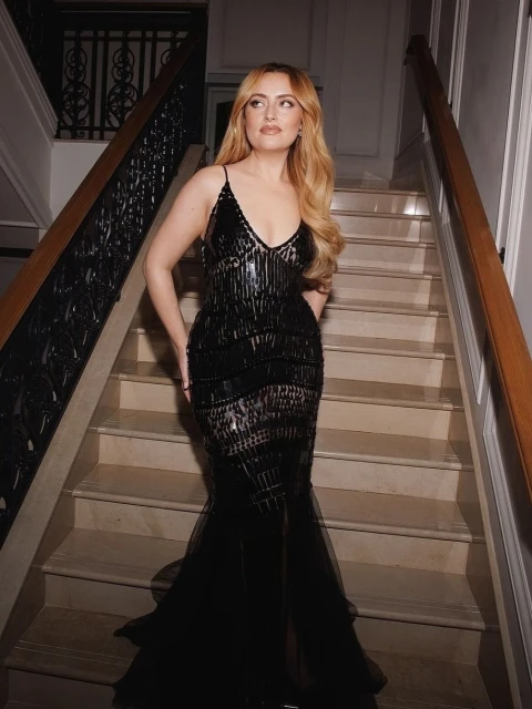 Woman with long, wavy blonde hair poses on marble stairs in a sequined black formal dress with a V-neck and fishtail skirt.